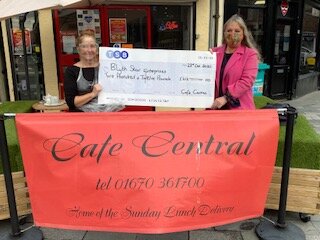 Women wearing PPE hold up a giant cheque in front of a cafe